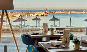 Utsikt från en restaurang mot en strand med parasoller och solstolar, samt en småbåtshamn med båtar. (AI-genererad text)