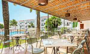 En utomhusrestaurang med bord och stolar under ett tak, med utsikt över en pool och hotellbyggnader i bakgrunden. (AI-genererad text)