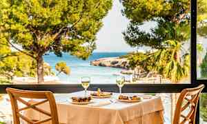 Ett dukat bord med mat och två glas vin, med utsikt mot havet genom ett stort fönster. (AI-genererad text)