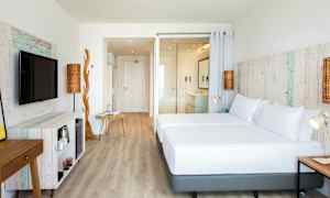 Ett ljust hotellrum med trädetaljer och moderna möbler. Rummet har en stor säng, en tv och en öppen planlösning. (AI-genererad text)