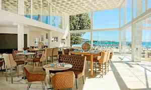 Ett modernt inrett restaurangutrymme med stora glasfönster som erbjuder utsikt mot en strand och havet utanför. (AI-genererad text)