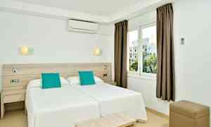 Ett hotellrum med två enkelsängar intill varandra, vit inredning och turkosa kuddar, samt ett fönster med utsikt. (AI-genererad text)
