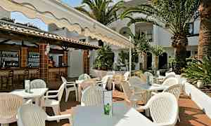 En uteservering med vita stolar och bord under en markis, omgiven av palmer och grönska. (AI-genererad text)