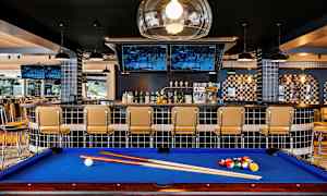 Biljardbord med blå duk framför en bar med stolar och tv-skärmar. (AI-genererad text)