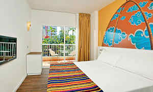 Ett hotellrum med en dubbelsäng och färgglad sängduk, en mönstrad väggmålning bakom sängen, ett fönster med utsikt över balkong och grönska. (AI-genererad text)