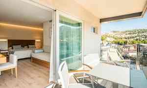 Ett hotellrum med två sängar och en sittmöbel, med en anslutande balkong där det finns bord och stolar samt utsikt över en grönskande kulle. (AI-genererad text)