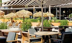 Ett utomhusområde med bord, stolar och solstolar under halmtak. (AI-genererad text)