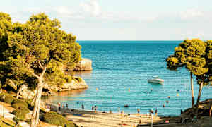 Strand med badgäster och träd vid ett klart blått hav, en båt är synlig på vattnet. (AI-genererad text)