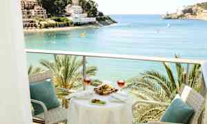 En balkong med bord och stolar, med utsikt över en vik med palmer och byggnader i bakgrunden. (AI-genererad text)