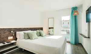 Ett hotellrum med en dubbelsäng, dekorativa kuddar, ett fönster med turkosa gardiner och en tv på väggen. (AI-genererad text)