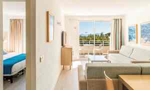 En ljus hotellsvit med ett vardagsrum, ett sovrum och en balkong med utsikt över träd och hav. (AI-genererad text)
