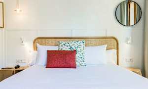Ett hotellrum med en bäddad dubbelsäng, prydd med vita kuddar och en röd dekorativ kudde framför en med blommönster. Sängens huvudgavel är i flätat trä. En rund spegel och vägglampor syns på en ljus bakgrund. (AI-genererad text)