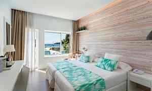 Ett ljust hotellrum med en stor säng, träpanel på väggen och ett fönster som vetter mot havet. (AI-genererad text)