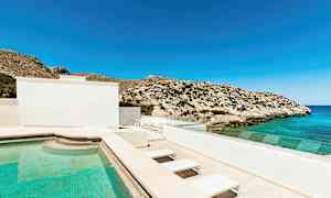 En utomhuspool vid ett hus, med utsikt över havet och klippiga berg under en klarblå himmel. (AI-genererad text)