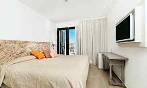 Ett hotellrum med en stor säng, dekorativa kuddar, en väggmonterad TV, och ett skrivbord. Balkongdörrar leder till en utsikt. (AI-genererad text)