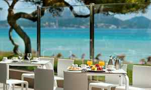 Bilden visar en terrass med vita bord och stolar, utsikt över blått hav och grönska. (AI-genererad text)