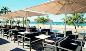 Ett utomhuskafé med bord, stolar och parasoller, med utsikt över en sandstrand och havet. (AI-genererad text)