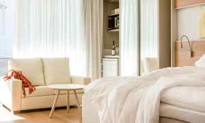 Ett ljust hotellrum med en soffa, ett litet bord och en bäddad säng. I bakgrunden finns ett minikök med kylskåp. (AI-genererad text)