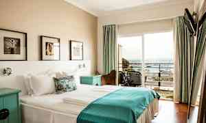 Ett hotellrum med en dubbelsäng och sängkläder i vitt och blått, väggkonst ovanför sängen och en balkong med utsikt över havet. (AI-genererad text)