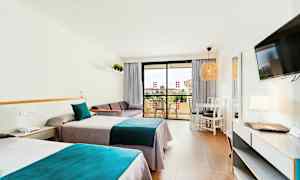 Ett hotellrum med två enkelsängar med blå och grå sängkläder, en soffa, ett bord med stolar och en balkong i bakgrunden. (AI-genererad text)