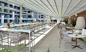 Ett modernt och ljust inrett terrass- eller restaurangområde med glasväggar, rader av bord och stolar samt dekorativa växter. (AI-genererad text)