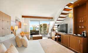 Ett hotellrum med trämöbler, en vit säng, en trappa och utsikt mot en terrass med hav i bakgrunden. (AI-genererad text)