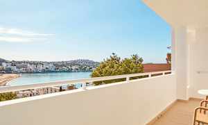 Utsikt från en balkong över en strand vid en vik, med byggnader och grönska i bakgrunden. (AI-genererad text)