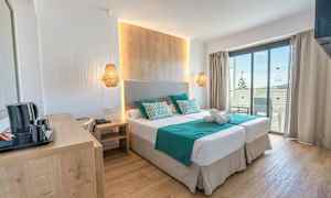 Ett hotellrum med två enkelsängar, en träpanel vid sängen, turkosa lakan och kuddar, samt en balkong med utsikt. (AI-genererad text)