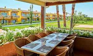 Bild på en utomhusterrass med ett dukat bord och stolar, med utsikt över en trädgård och gula byggnader i bakgrunden. (AI-genererad text)