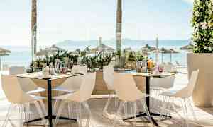 Bild av en utomhusrestaurang med vita stolar och bord, med havsutsikt och parasoller i bakgrunden. (AI-genererad text)