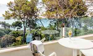 En balkong med utsikt över träd och hav. På balkongen står ett vitt bord med ett glas champagne och en ställning med en kylhink för en champagneflaska. (AI-genererad text)