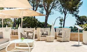 Sittgrupp utomhus med vita möbler av flätad design, beige kuddar, och parasoller. Gröna träd och havet i bakgrunden. (AI-genererad text)