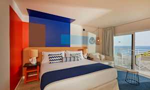 Ett rum med en dubbelsäng, dekorativt målade väggar, sittmöbler och en utsikt mot havet genom stora fönster. (AI-genererad text)