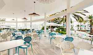 Utomhusterrass med bord och stolar, omgiven av palmer och vita byggnader. (AI-genererad text)