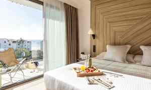 Ett ljust hotellrum med en bäddad säng med en matbricka. Stora glasdörrar leder till en balkong med en utsikt över havet. (AI-genererad text)