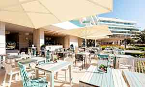 En utomhusrestaurang med bord och parasoller, omgivande byggnader syns i bakgrunden. (AI-genererad text)