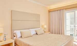 Rum med en dubbelsäng, beige sängkläder och lampa på nattygsbordet vid sidan av fönster med gardiner. (AI-genererad text)