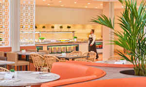 Bild på en matsal med moderna möbler, där bord med stolar står placerade. En buffé med olika maträtter syns i bakgrunden. (AI-genererad text)