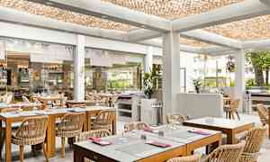 En ljus restaurang med träbord och rottingstolar, placerad i en modern inomhusmiljö med ett dekorativt tak och glasväggar som öppnar upp mot utsidan. (AI-genererad text)