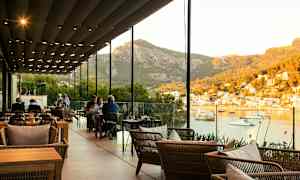 En restaurang med sittplatser utomhus under ett täcke med utsikt över en hamn och berg i bakgrunden. (AI-genererad text)