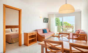 En hotellrumsvit med ett bord, stolar, soffa och ett sovrum synligt genom en öppen dörr. (AI-genererad text)