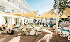 En utomhusterrass vid en byggnad, med bord, stolar och skugggivande markiser. (AI-genererad text)