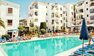 Ett poolområde med solstolar runt en klarblå pool, framför en vit byggnad med balkonger. (AI-genererad text)