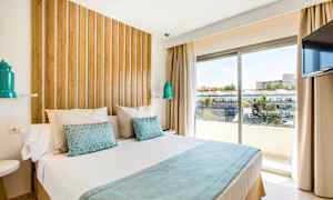 Ett modernt hotellrum med en stor säng, träpanel bakom sängen, vita och blå sängkläder, samt en fönstervy över byggnader och träd utanför. (AI-genererad text)