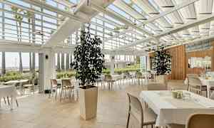 Restaurang med bord och stolar under ett glastak, omgiven av gröna växter och palmer. (AI-genererad text)