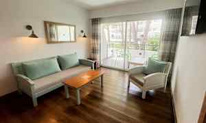 En sittgrupp med soffbord och stolar, samt ett glasfönster som leder till en balkong. (AI-genererad text)