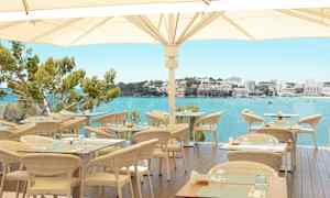 En utomhusrestaurang med rottingmöbler och utsikt över en klarblå havsbukt och byggnader i bakgrunden. (AI-genererad text)