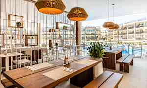 Restaurang med träbord och ljusa stolar, dekorerad med växter och lampor, med utsikt mot en pool och hotellbyggnader. (AI-genererad text)