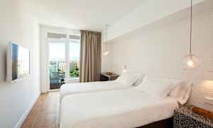 Ett hotellrum med två enkelsängar, vita sängkläder, en vägg-TV och utsikt över en stad genom glasdörrar. (AI-genererad text)