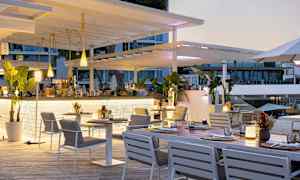 En utomhusrestaurang med vita möbler och gröna växter, belägen nära en byggnad med glasfönster. (AI-genererad text)